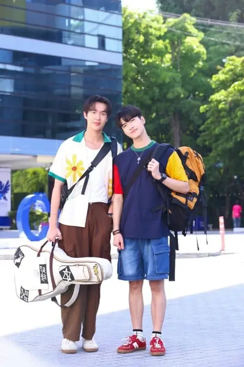 unlucky-bae-the-series-2026-hinh-anh-7 Unlucky Bae The Series hai nam diễn viên mang balo và túi đàn guitar đứng chụp ảnh trước tòa nhà