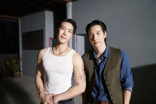 lovers-and-gangsters-the-series-2026-hinh-anh-8 Lovers and Gangsters The Series – hai diễn viên nam đứng cạnh nhau tại hậu trường, một người mặc áo ba lỗ trắng để lộ hình xăm, một người mặc vest xanh